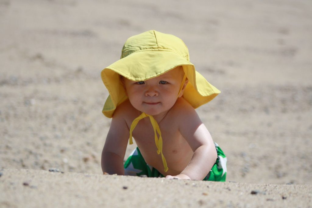 A young boy in a yellow beach hat crawls across the sand on Cape Cod.